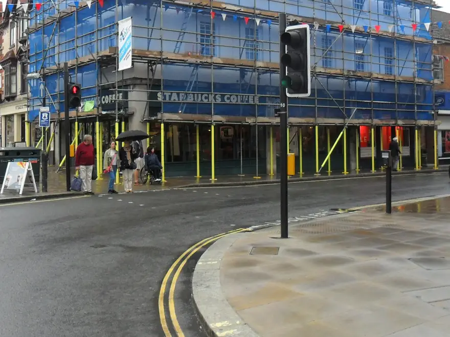 Pedestrians walking beneath a netted scaffold on Henley high street, with the shopfronts clear