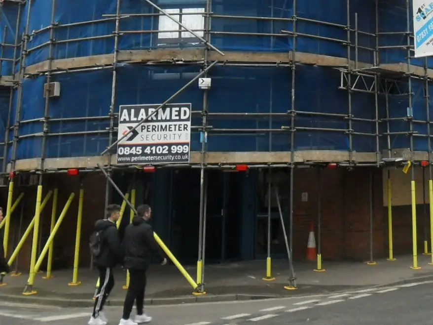 Base of a netted office-block scaffold in Maidenhead with an alarm sign and yellow upright protection